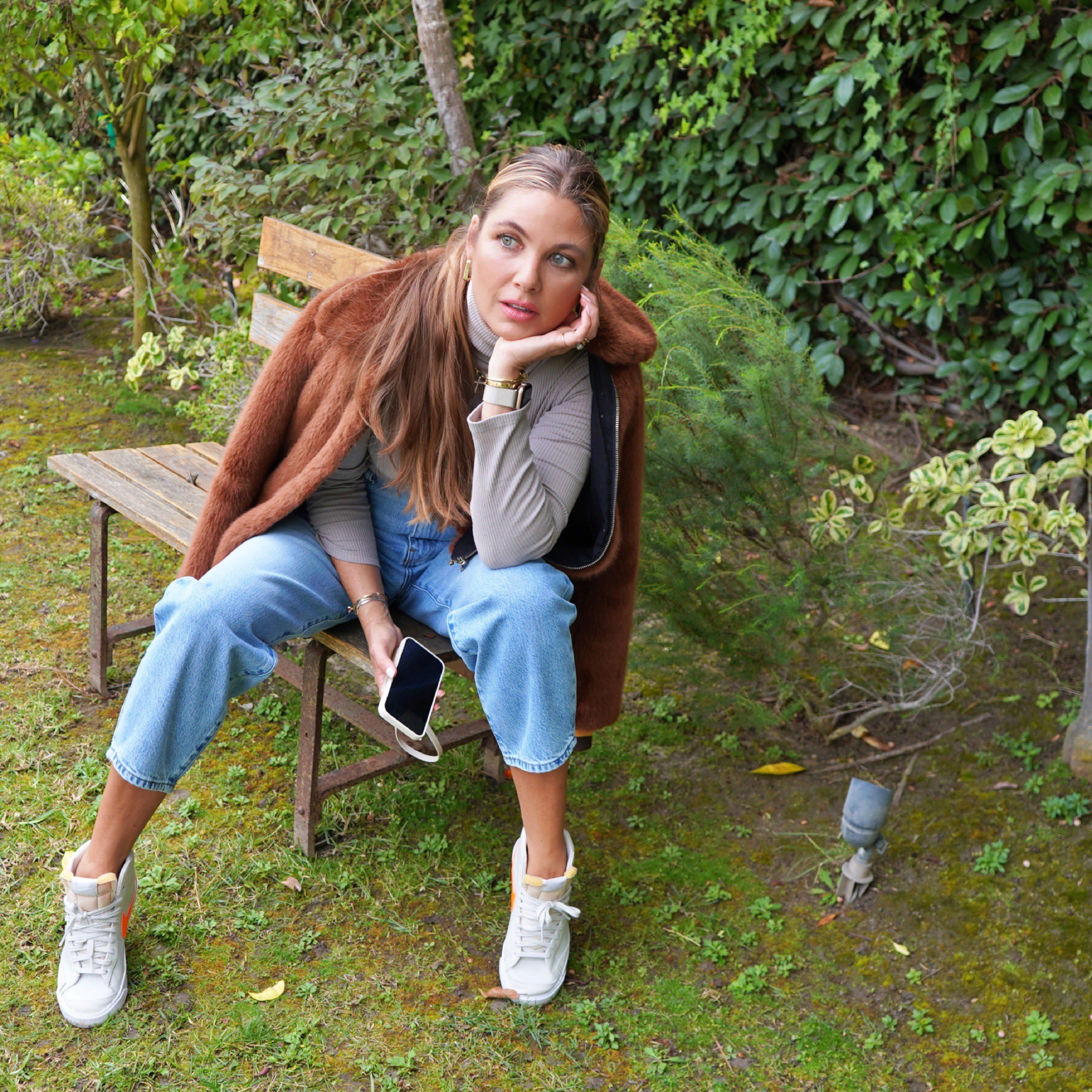 woman sitting on a bench and wearing pants, shirt, and brown jacket 